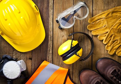 Personal safety workwear items shot directly above. The safety equipment includes a hard hat, goggles, gloves, steel toe shoes, earmuff a respiratory mask and a safety vest. The background is a rustic wood table scratched and stained with vertical stripes.  Predominant colors are yellow and brown. DSRL studio photo taken with Canon EOS 5D Mk II